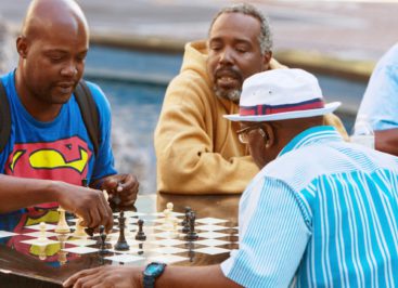 African-American men playing a friendly game of chess at an outdoor park, while others watch