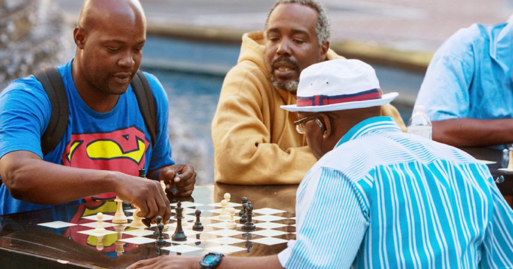 African-American men playing a friendly game of chess at an outdoor park, while others watch